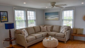A living room with a beige sectional sofa, a round coffee table, and a ceiling fan. There are two windows with blinds, a lamp, and a painting on the wall.