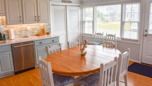 A kitchen with white cabinets, a wooden dining table, and a view through the windows.
