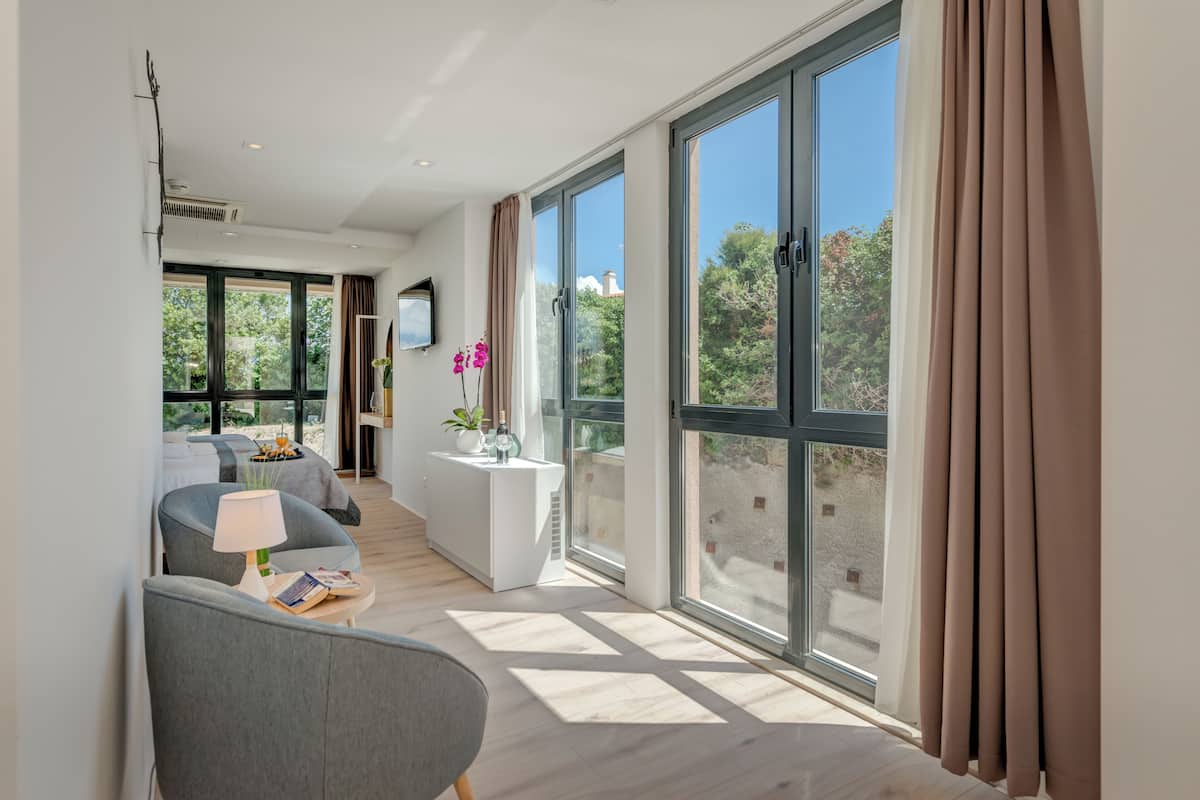 A modern hotel room with a large sliding glass door, a sofa, a small table with a lamp, and a view of greenery outside.