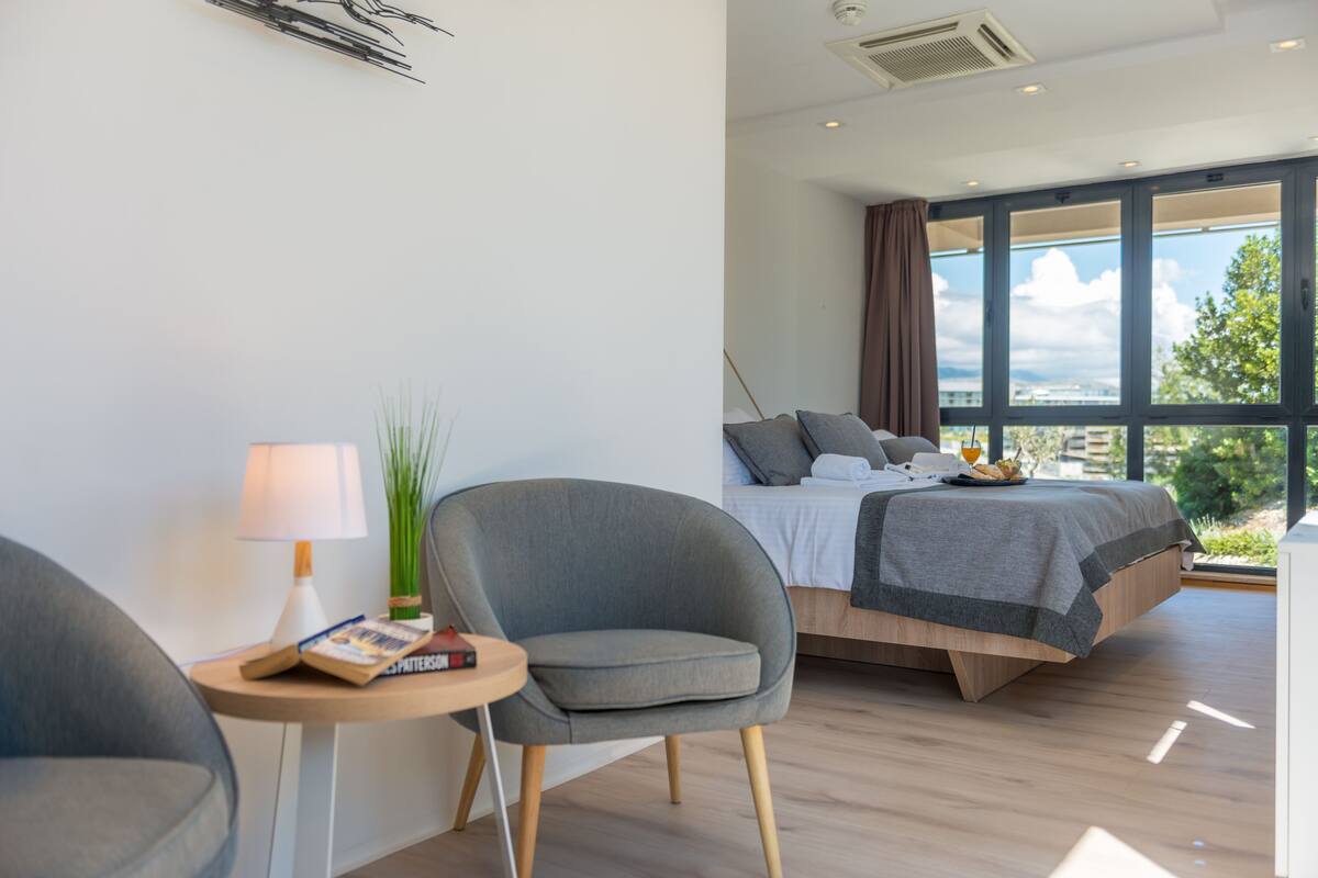 A modern bedroom with a bed, a chair, a small table with a lamp, and a view of the outdoors.