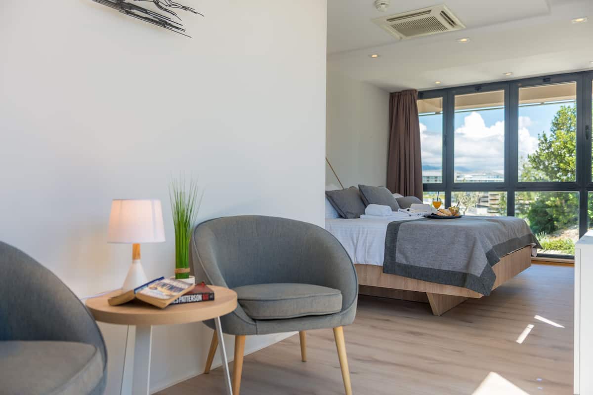 A modern bedroom with a bed, a chair, a small table with a lamp, and a view of the outdoors.
