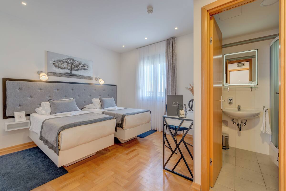 A hotel room with two beds, a bedside table, a nightstand, a small table with a lamp, a mirror, and a bathroom with a sink and mirror.