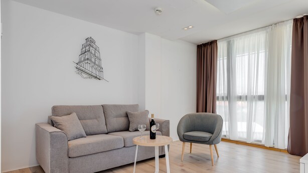 A modern living room with a grey sofa, a small round table with a bottle and glasses, a grey armchair, and a wall-mounted artwork of a building. - PAX (Split)