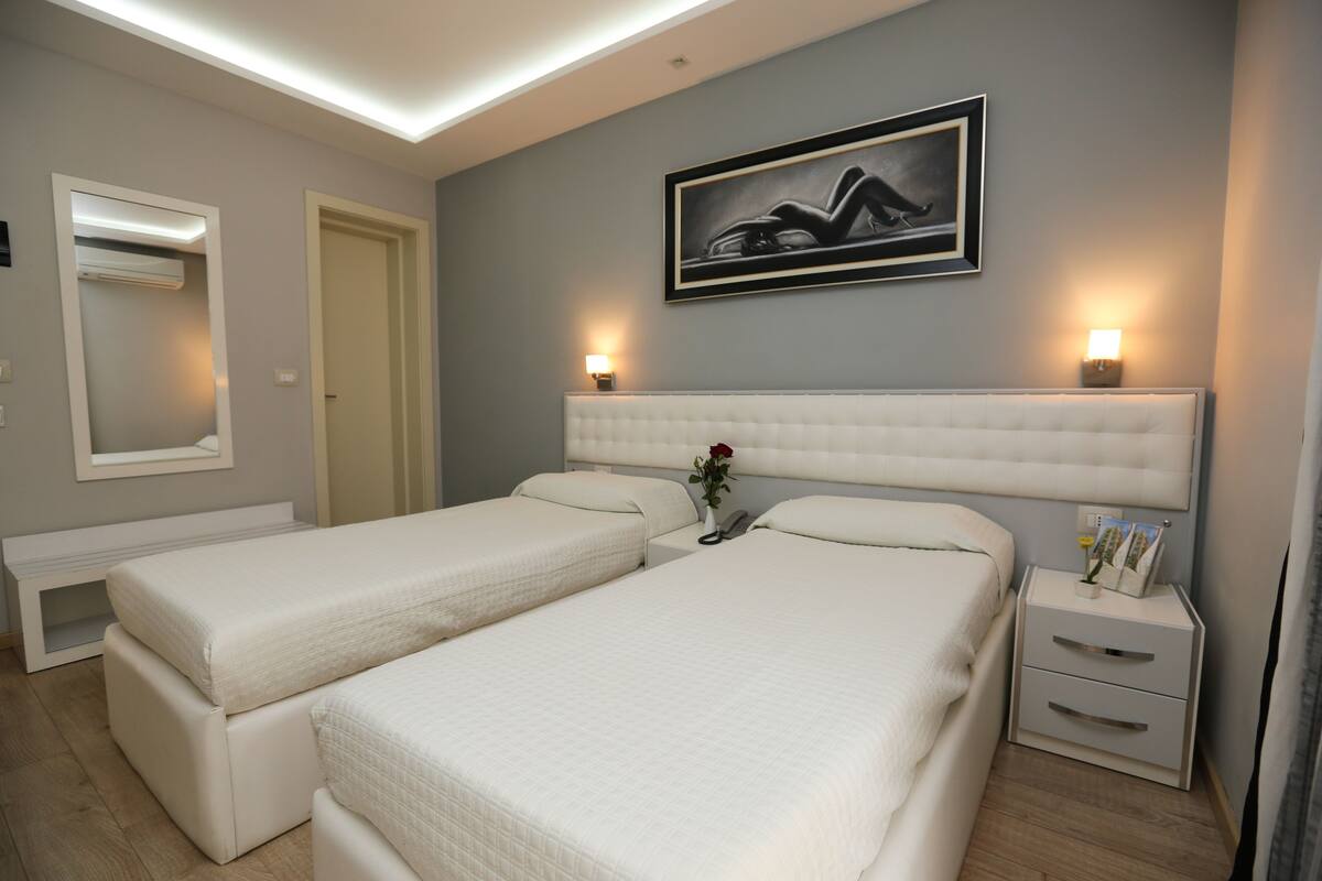 A hotel room with two beds, a bedside table with a lamp, a framed picture on the wall, and a mirror above the door.
