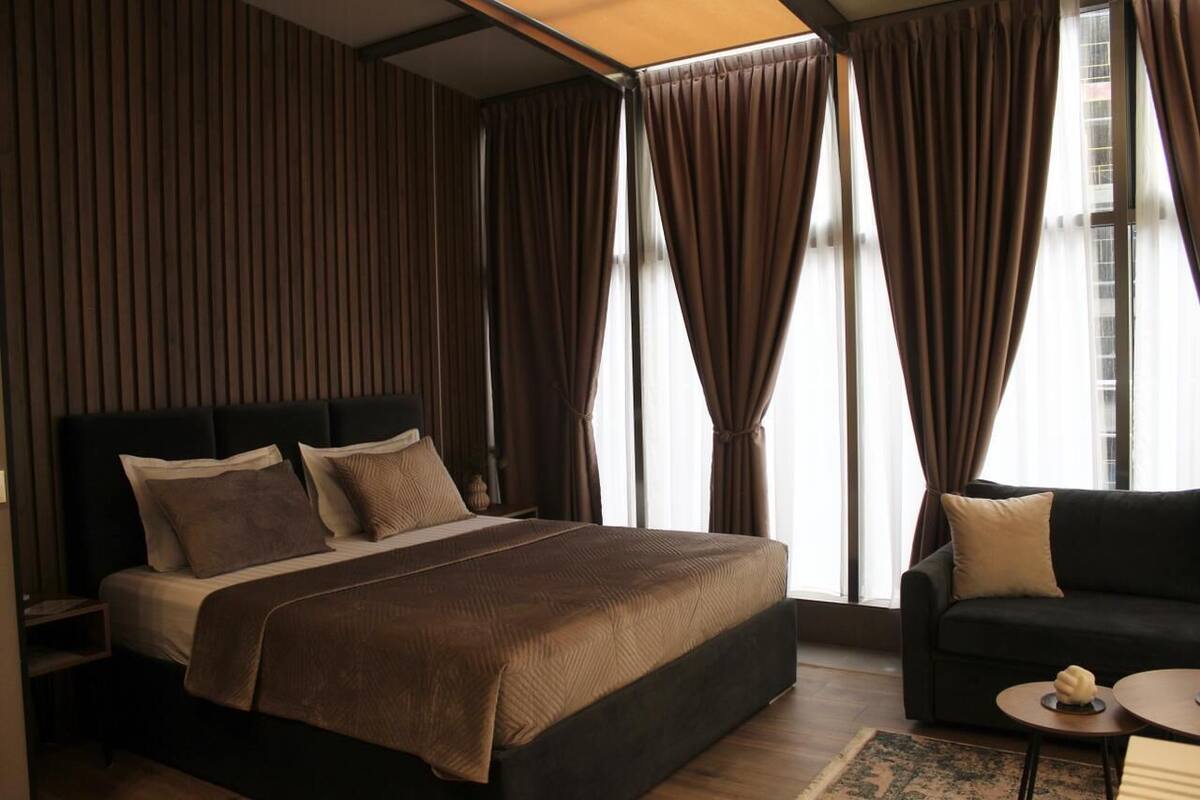 A modern bedroom with a large bed, dark headboard, and curtains over windows.