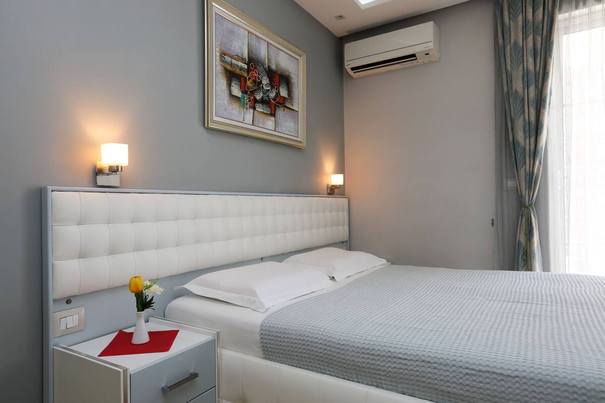 A hotel room with a large bed, two bedside tables with lamps, a painting on the wall, and an air conditioning unit.
