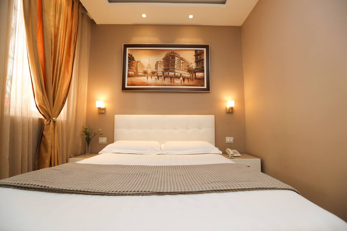 A hotel room with a large bed, a headboard, two bedside lamps, a nightstand, a framed picture on the wall, and a curtain.
