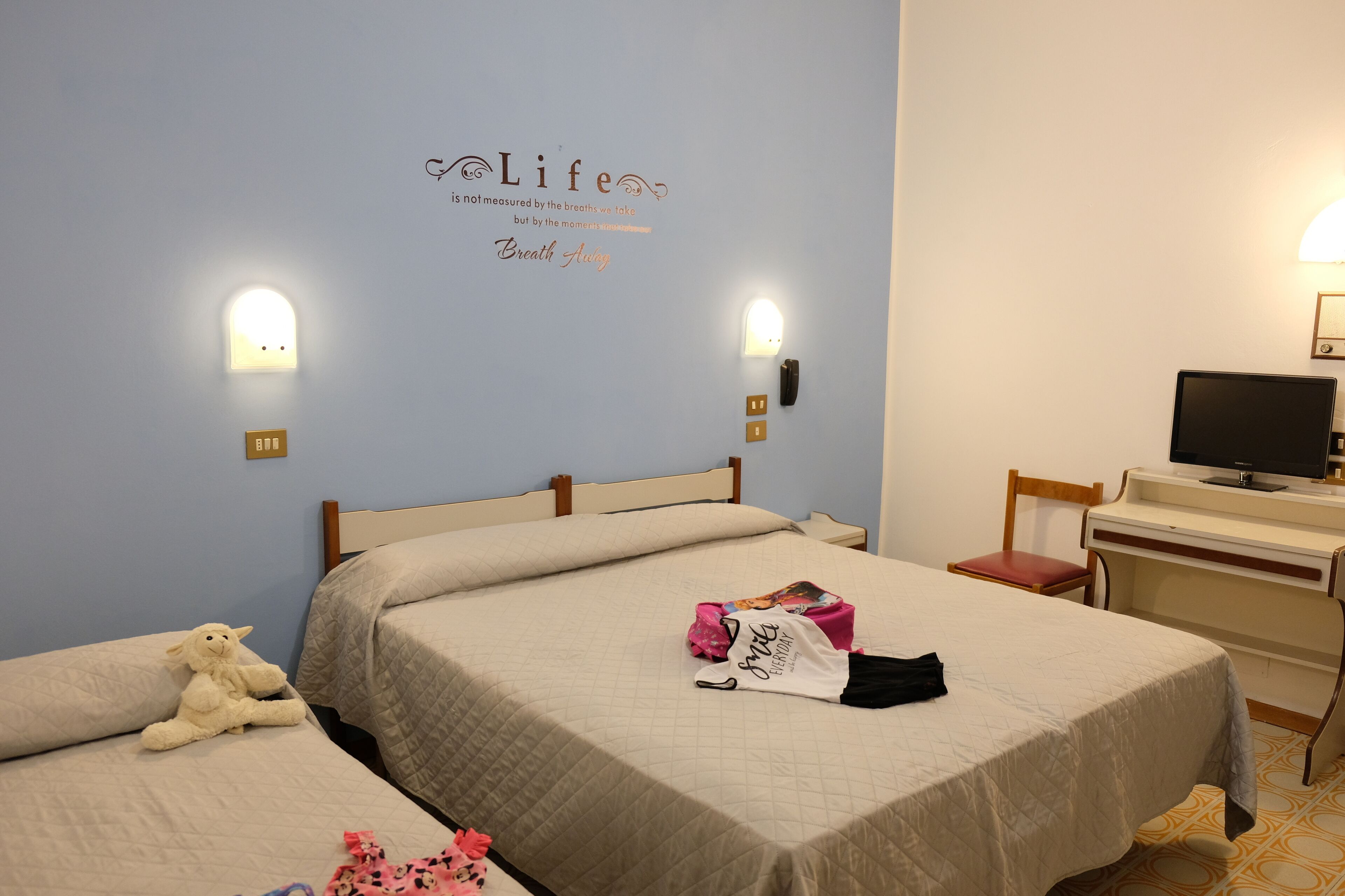 Une chambre d’hôtel avec deux lits, un bureau, une chaise, une télévision et un mur orné d’une citation inspirante.
