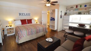 Un dormitorio con cama, mesitas de noche, cómoda, ventilador de techo y una zona de cocina con taburetes y estanterías.