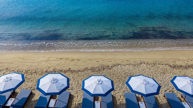 A beach with clear blue water, sandy shore, and several blue umbrellas and lounge chairs. - Aegon Mykonos, Autograph Collection (Mykonos)