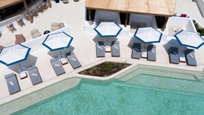 A pool area with lounge chairs and umbrellas, a small garden, and a white building in the background. - Aegon Mykonos, Autograph Collection (Mykonos)