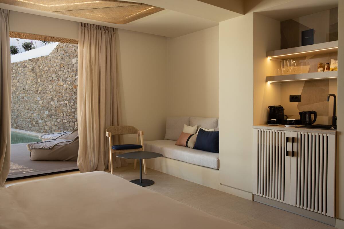 A modern hotel room with a bed, a small table, a chair, a sofa with cushions, a kitchen area with a coffee machine, and a view of a stone wall and greenery outside.