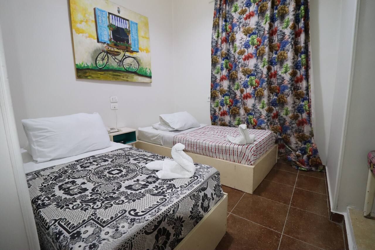 A room with two beds, one with a patterned bedspread and the other with a striped one. There is a painting on the wall above the beds, and a colorful curtain on the window.