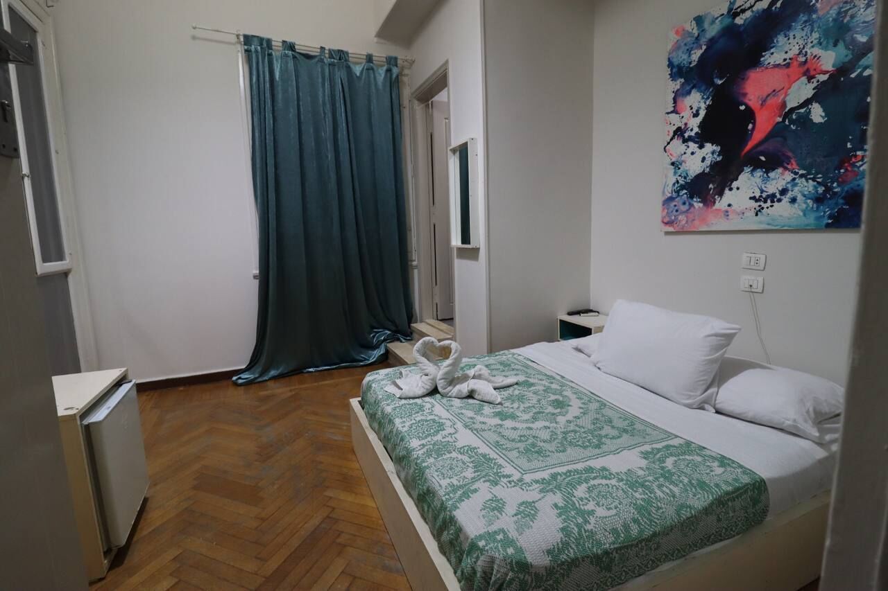 A bedroom with a bed, a green and white patterned bedspread, a white pillow, a wall painting, a window with a curtain, and a radiator.