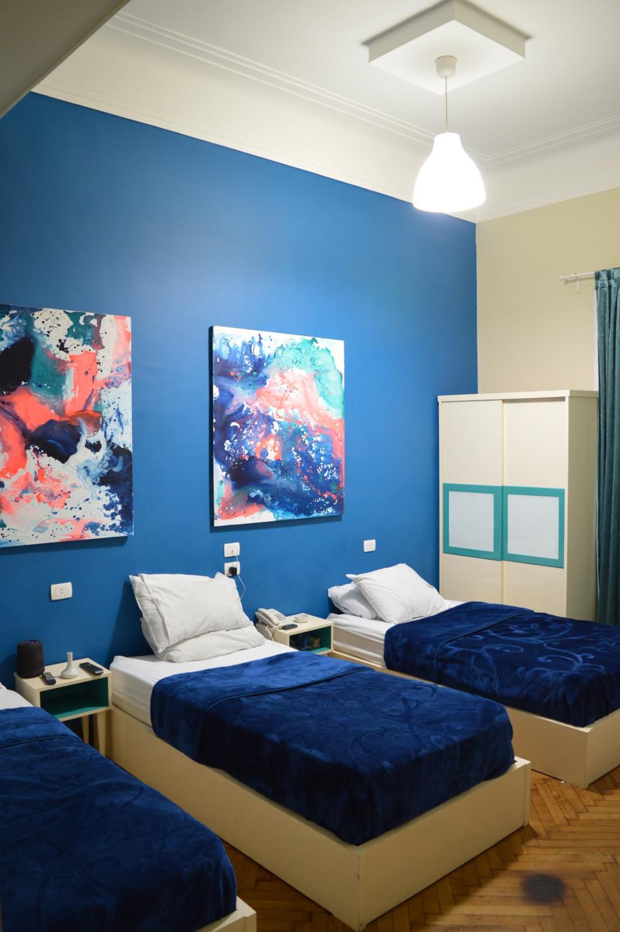 A room with three beds, blue walls, abstract paintings, a wardrobe, and a hanging light fixture.