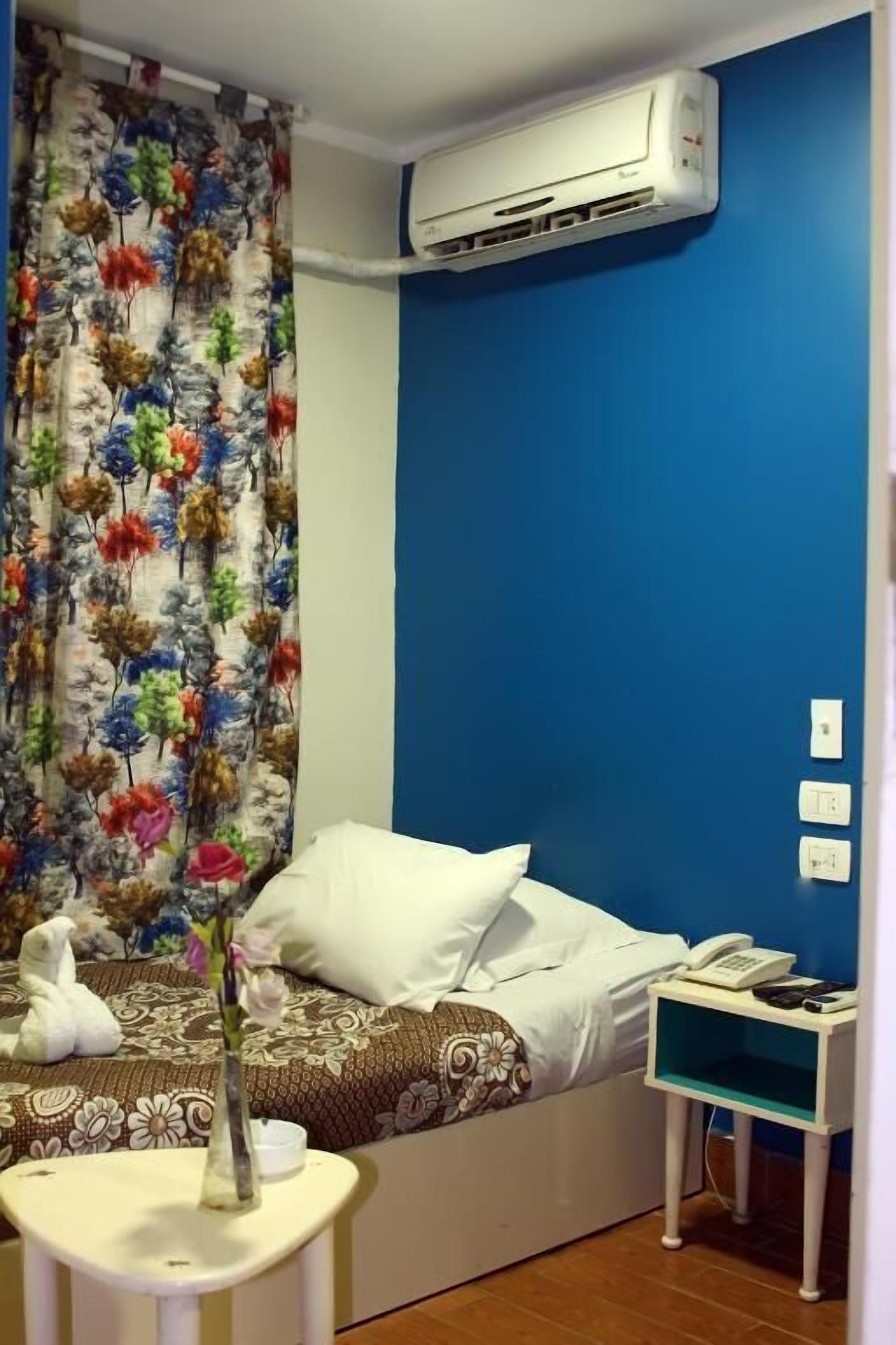 A hotel room with a bed, a nightstand, an air conditioner, and a curtain with a floral pattern.