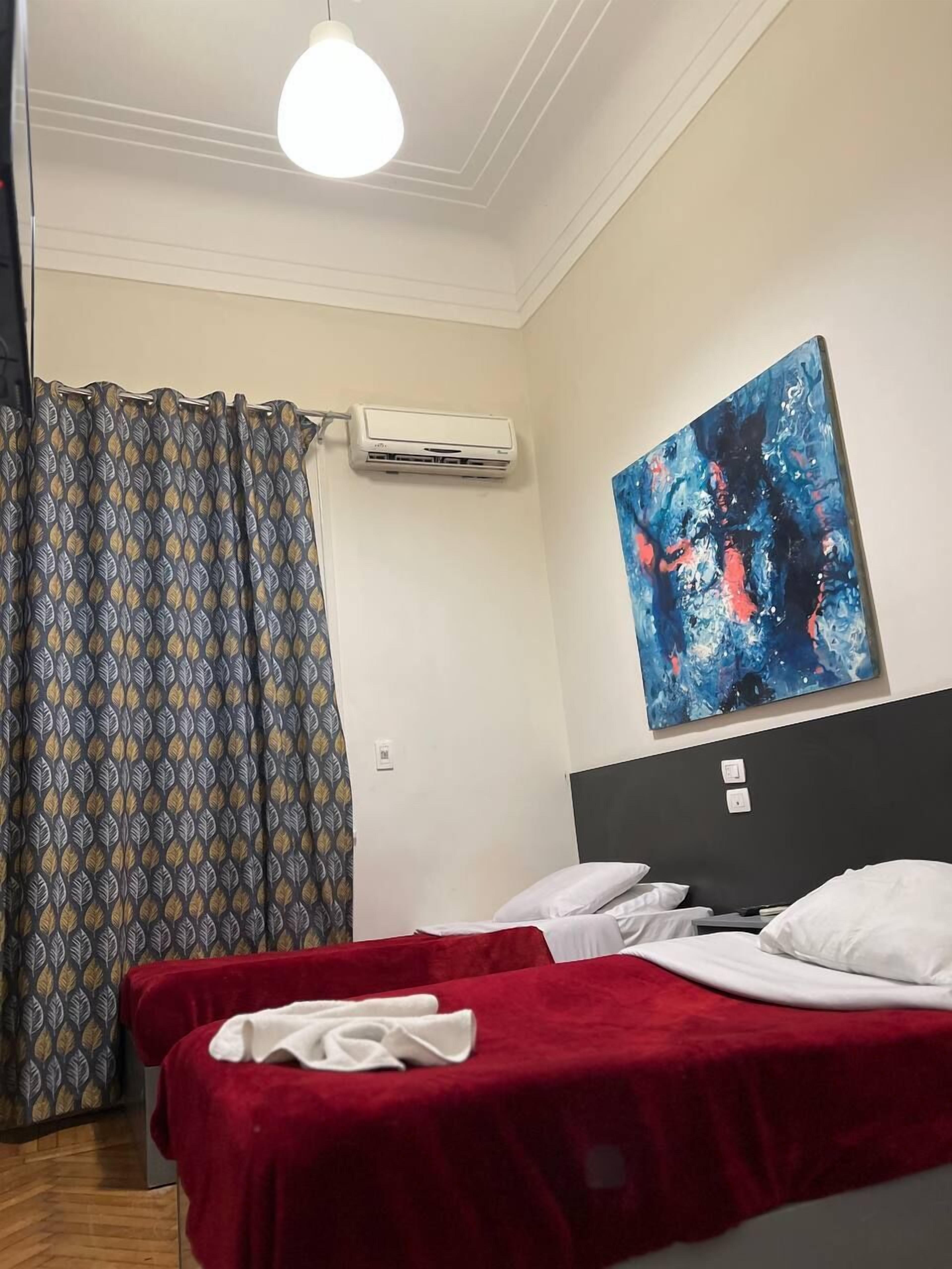 A hotel room with a bed, a red blanket, an air conditioning unit, a patterned curtain, and a painting on the wall.