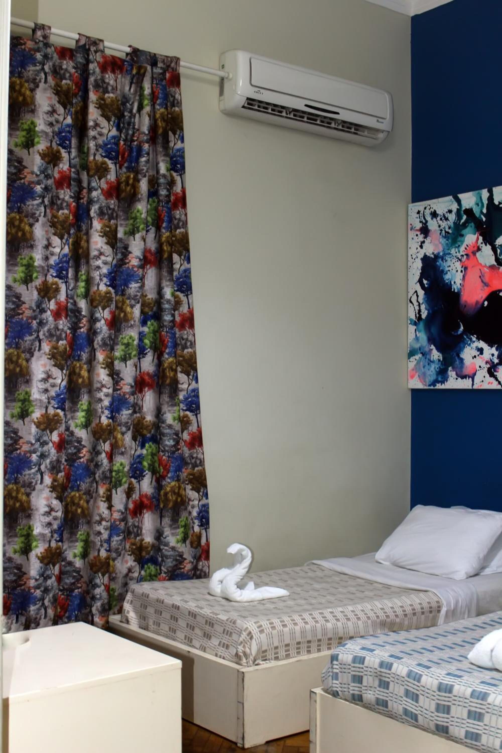 Ein Hotelzimmer mit zwei Betten, einer weißen Klimaanlage, einem farbenfrohen Vorhang und einem Bild an der Wand.
