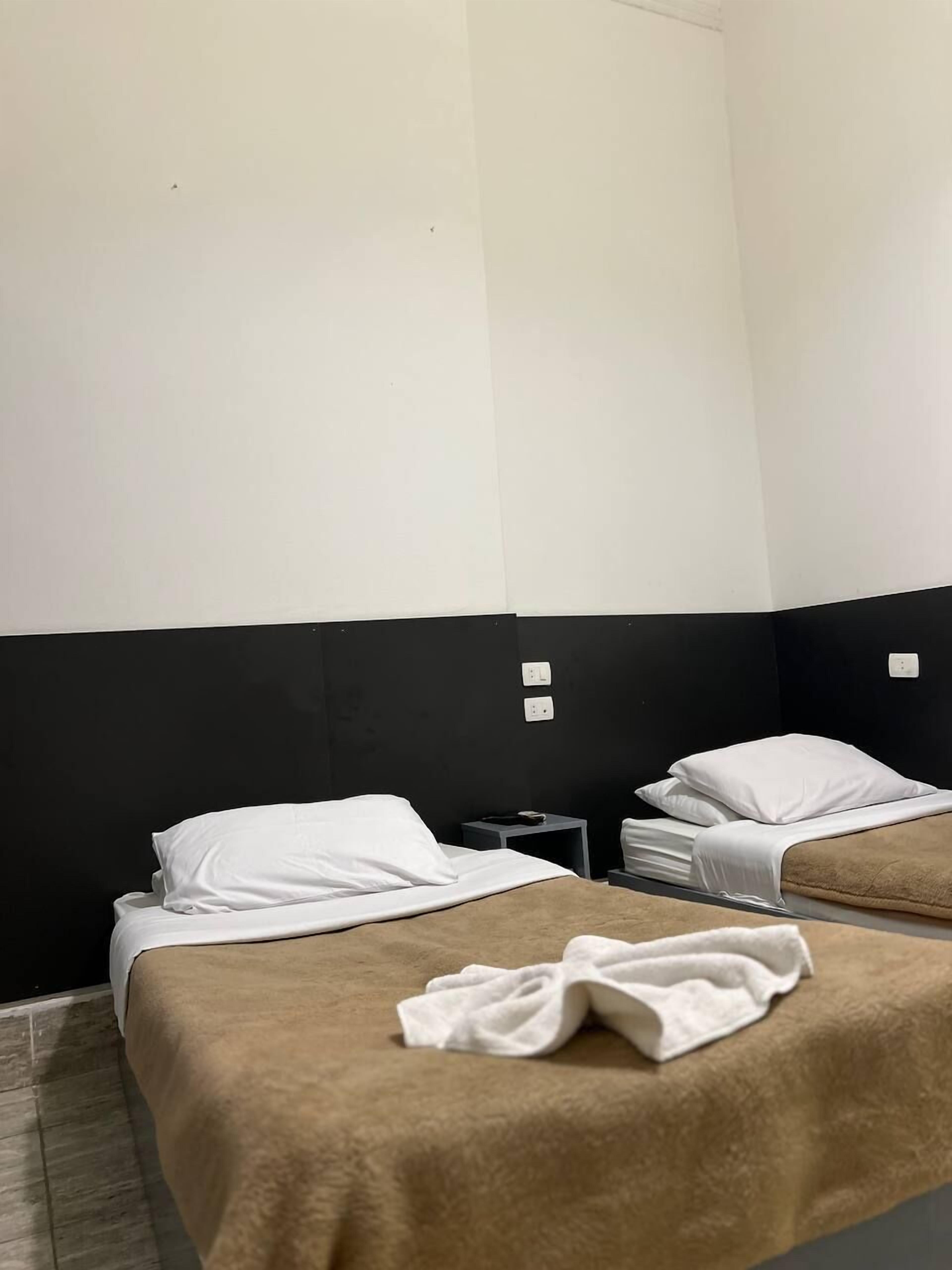 A hotel room with two beds, each with a white pillow and a brown blanket. There is a small table between the beds with a remote control on it. The walls are painted black and white.