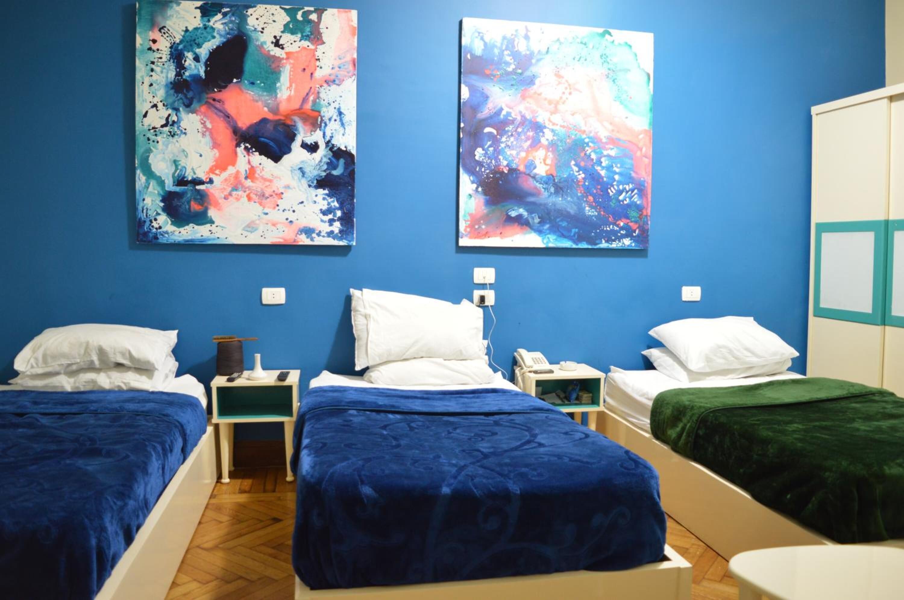 A hotel room with two beds, each with a blue bedspread, white pillows, and a bedside table with a lamp and phone. There are abstract paintings on the wall and a wardrobe with a mirror.