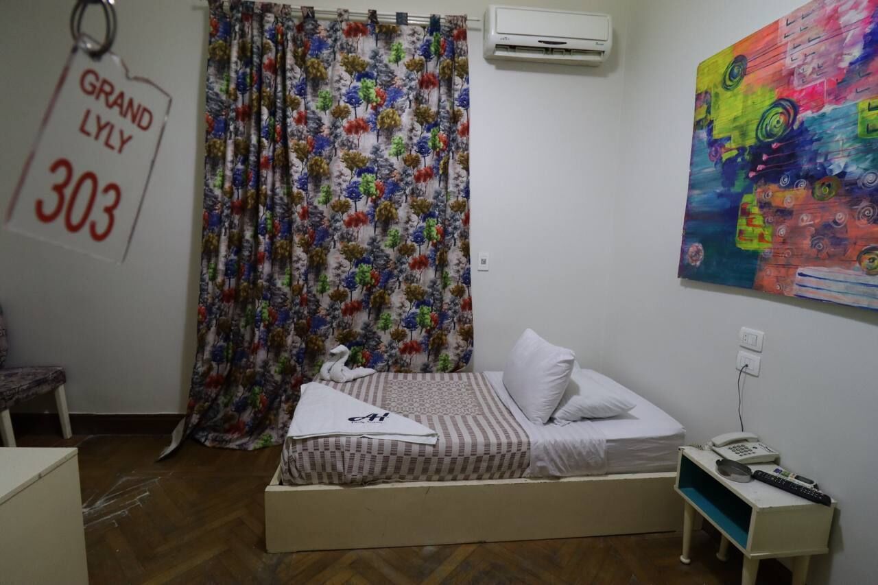 Ein Schlafzimmer mit einem Bett, einem weißen Kopfkissen, einem Nachttisch mit Telefon und Fernbedienung, einem farbenfrohen Gemälde an der Wand und einem blumigen Vorhang.
