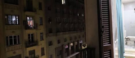 A nighttime view of a building with multiple floors and balconies, a partially open door revealing a room with a toilet and a blue curtain, and a railing along the balcony.
