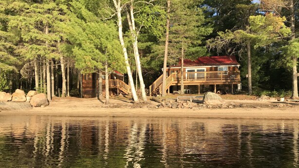 A lakeside cabin with a wooden deck and stairs leading to a red-roofed main building. - Loon Lodge - Golden Lake (Cottage & Bunkie -Sleeps 8 ) (Killaloe)