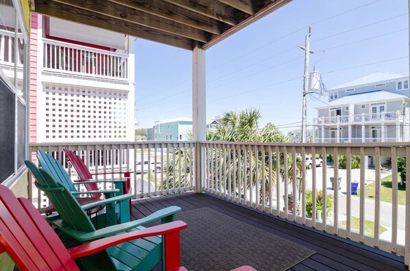 A covered porch with red and green Adirondack chairs, a white fence, and a view of the street and neighboring buildings.