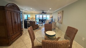 A spacious living room with a glass dining table surrounded by wicker chairs, a large wooden cabinet, and a view of the outdoors through large windows.