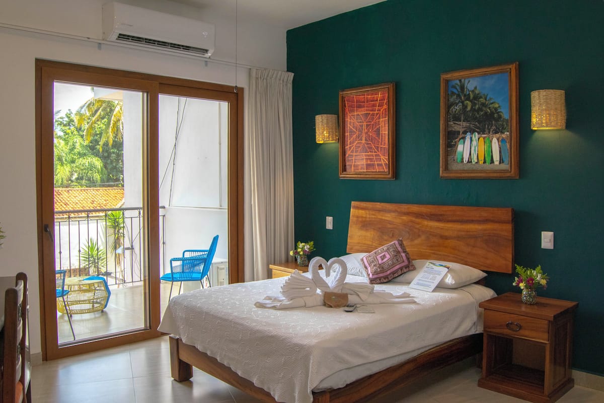 Habitación de hotel con cama, mesita de noche, cabecero de madera, ventana con vista al exterior y pared con dos cuadros enmarcados.