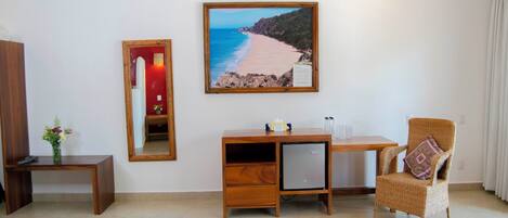 Habitación de hotel con un escritorio de madera, una silla, un refrigerador pequeño, un espejo y un cuadro con vista al mar.
