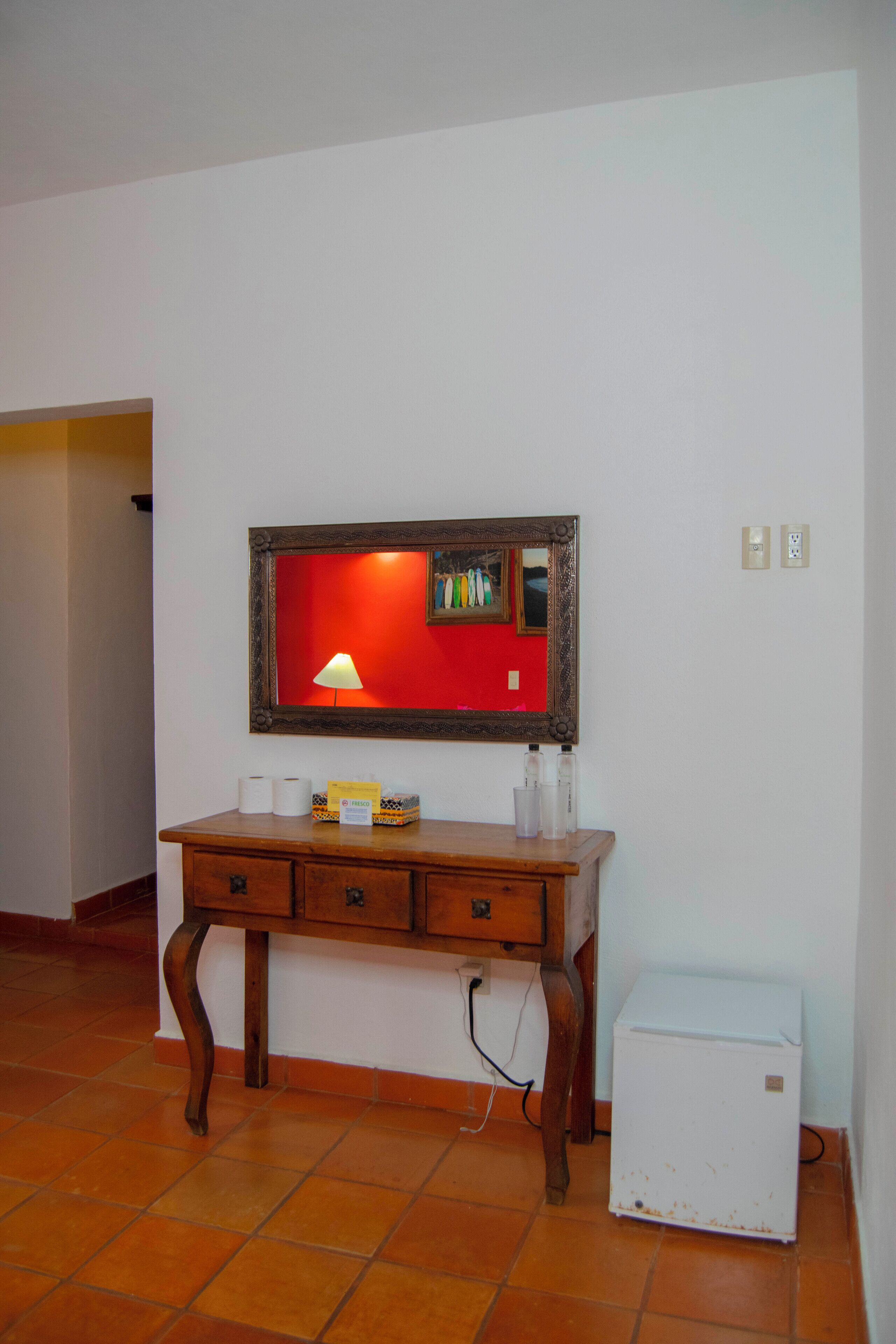 Une pièce avec un bureau en bois, un petit réfrigérateur blanc et un miroir avec un cadre rouge.