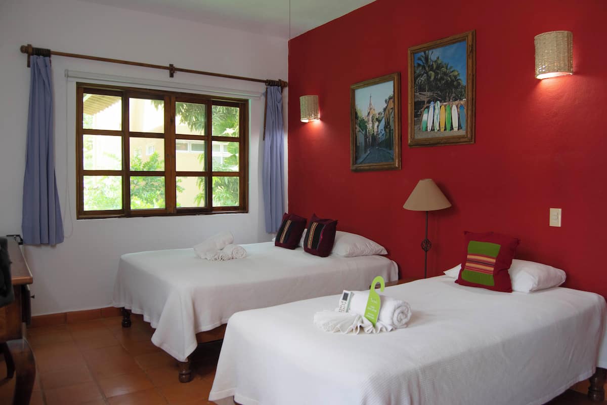 Habitación de hotel con dos camas, un ventanal con cortinas, un cuadro y una lámpara.