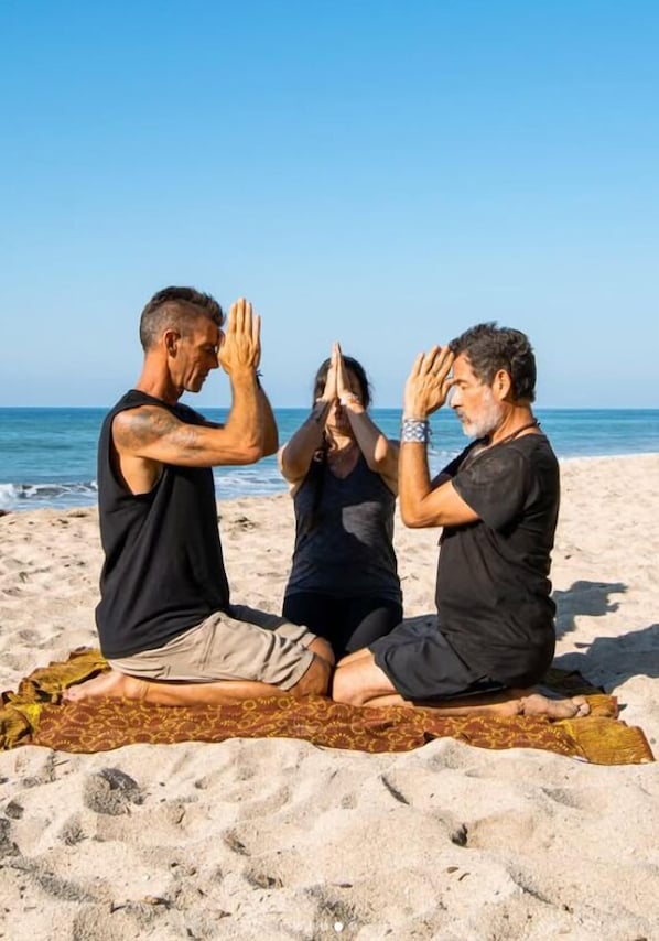 Três pessoas sentadas em uma esteira na praia, praticando uma pose de yoga.