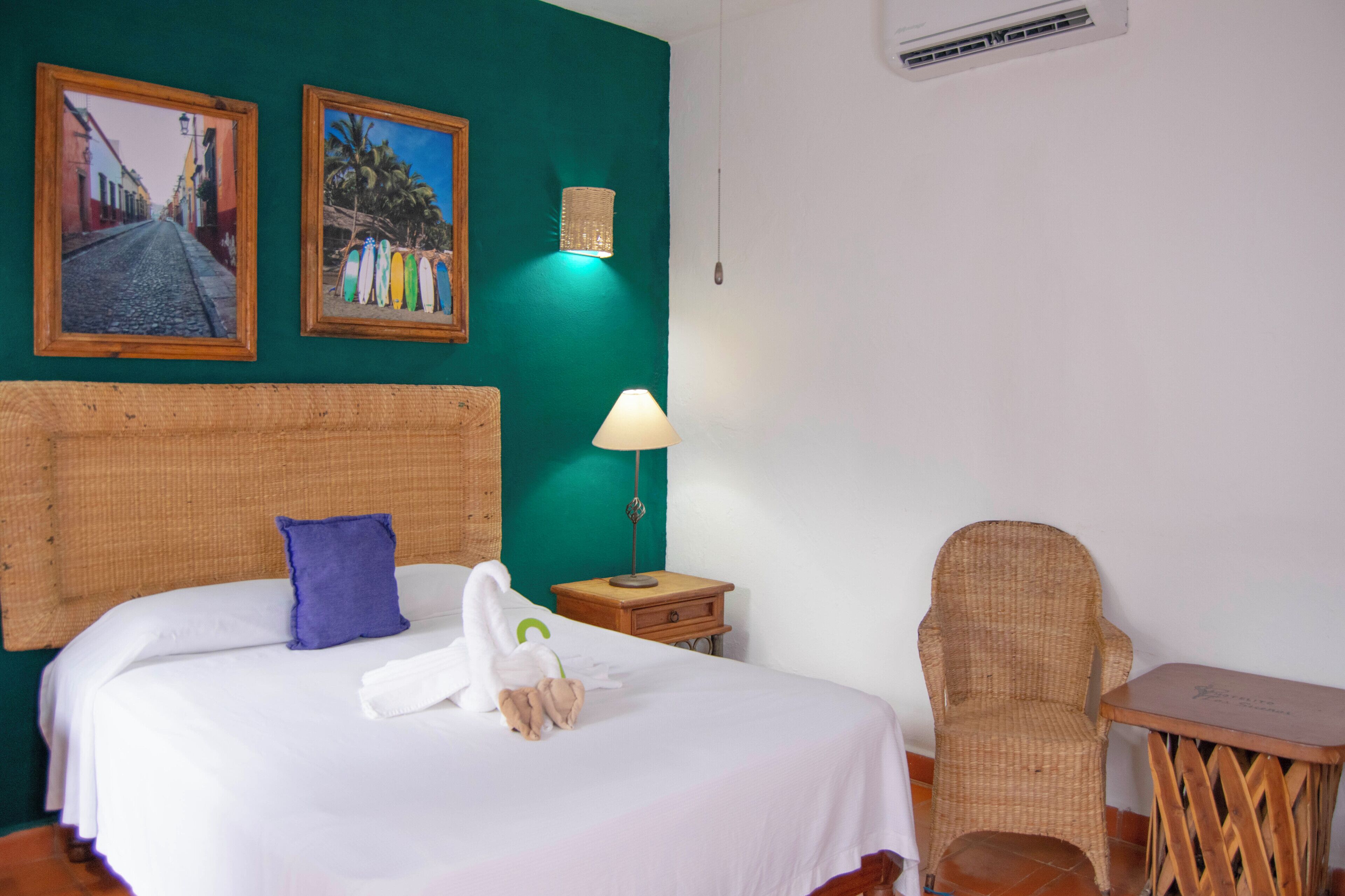 A hotel room with a bed, a chair, a small table, and two framed pictures on the wall.