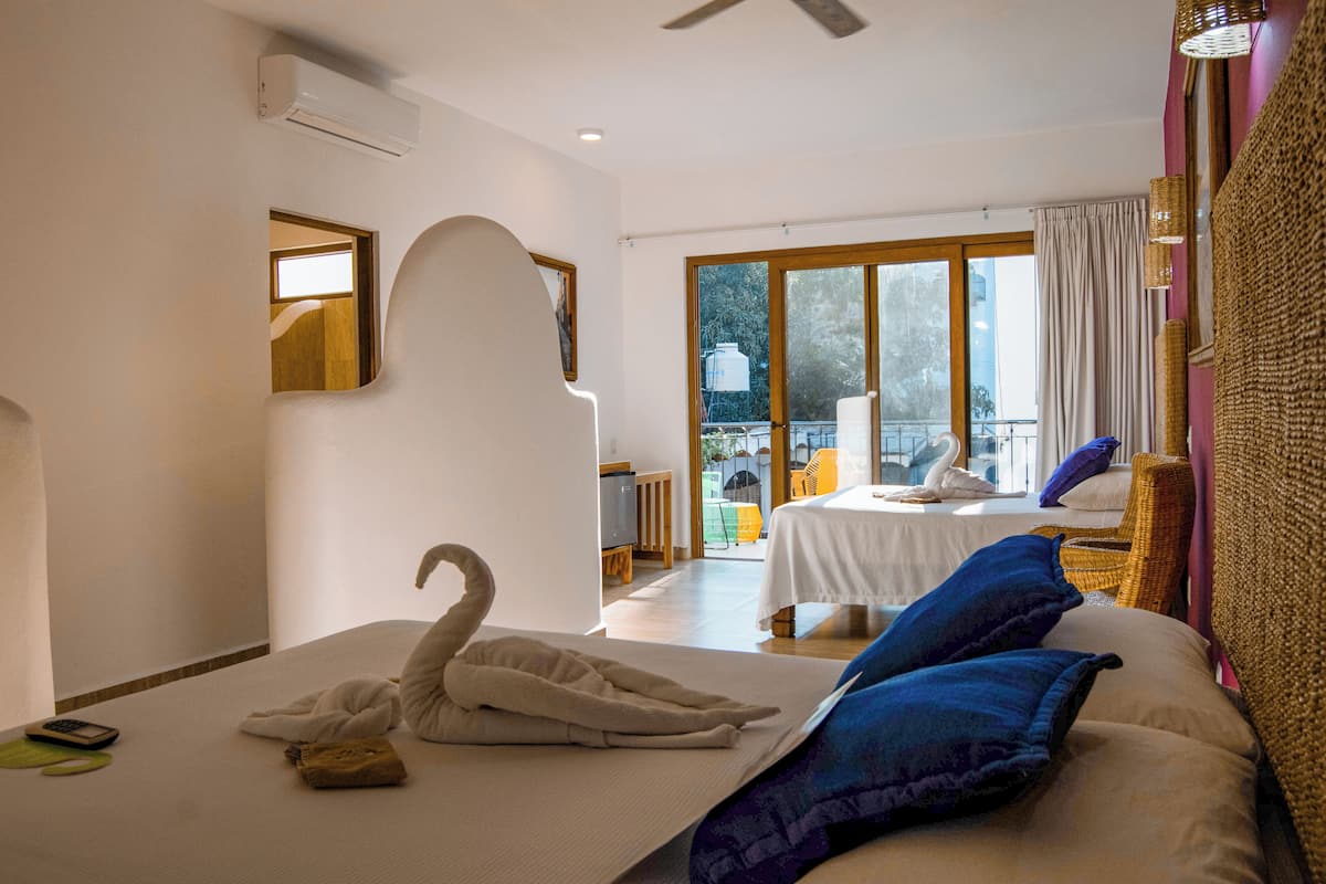 Habitación de hotel con una cama grande, un cojín con forma de cisne, una segunda cama al fondo y un balcón con vistas.