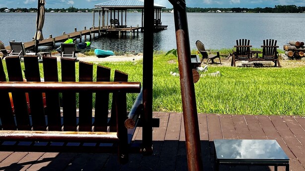 A wooden deck with a covered seating area overlooking a lake, a dock, and a grassy area with chairs. - Lake Front Sebring Villa , Relax- Quiet with Breathtaking views! (Sebring)