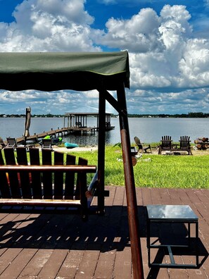 A wooden deck with a covered seating area overlooking a lake, a dock, and a grassy area with chairs. - Lake Front Sebring Villa , Relax- Quiet with Breathtaking views! (Sebring)