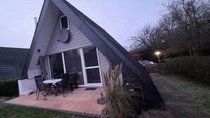 A house with a sloped roof, a patio with a table and chairs, and a large window. - Holiday house at Wittensee (approx. 2 minutes walk to the lake) with small Rowing boat, from April 2019 (Bünsdorf)