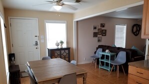 Un coin repas avec une table et des chaises en bois, un ventilateur de plafond et une vue sur un salon comprenant un canapé et une table basse.