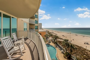 A balcony with a pool, beach view, and lounge chairs.