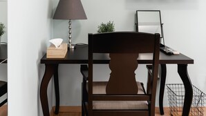 Uma sala de estar moderna com uma escrivaninha de madeira escura, uma cadeira, uma televisão fixada na parede e um pequeno estante com objetos decorativos.