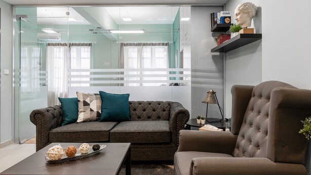 Uma sala de estar moderna com um sofá cinza, uma poltrona marrom e uma mesa de centro com bolas decorativas. Há uma estante com livros e um busto na parede.
