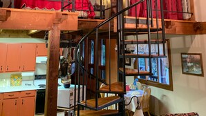 Un interior rústico con una escalera de madera, un dormitorio en entrepiso con cortinas rojas y una zona de cocina.
