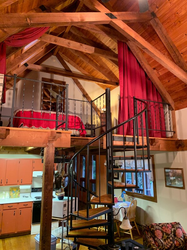Cozy loft space with a red bed, kitchen, and dining area, featuring exposed beams and a vaulted ceiling.
