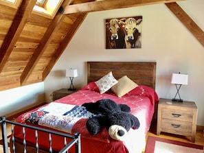 Un dormitorio con techo de madera, una cama con colcha roja, dos mesitas de noche con lámparas y un cuadro de vacas en la pared.