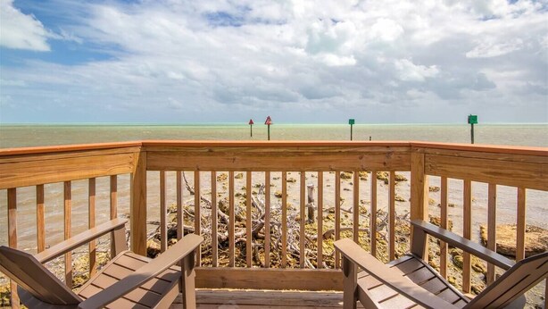 Eine Holzterrasse mit zwei Stühlen, die auf einen Strand ausgerichtet sind, mit einem überdachten Reetdach.