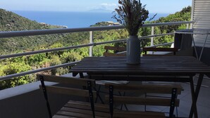 Un balcón con una mesa y sillas, un jarrón con flores secas y una vista panorámica del mar y las colinas.