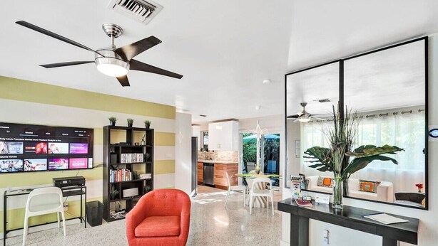 A modern living room with a TV, bookshelves, a red armchair, a dining area, and a kitchen in the background. - Heated pool, parking, 5 min walk to food and fun! (Wilton Manors)