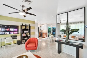 Un salon moderne avec un téléviseur, des étagères, un fauteuil rouge, un coin repas et une cuisine en arrière-plan.